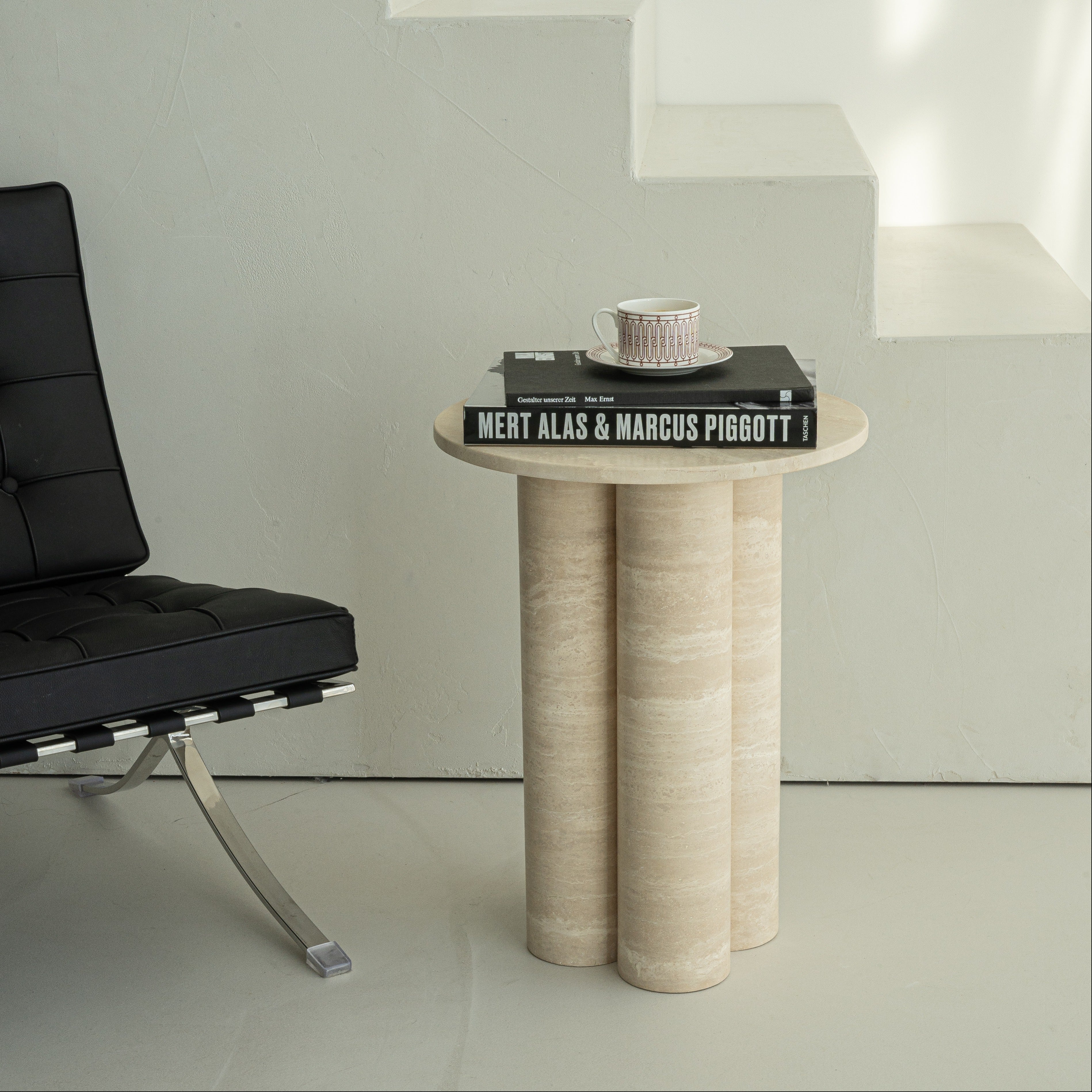 Klassischer Beistelltisch aus Travertin mit runder Tischplatte und drei zylinderförmigen Beinen, dekoriert mit einem Buch und einer eleganten Tasse, inszeniert in einem minimalistischen Wohnambiente mit schwarzem Ledersessel und Treppenstruktur.