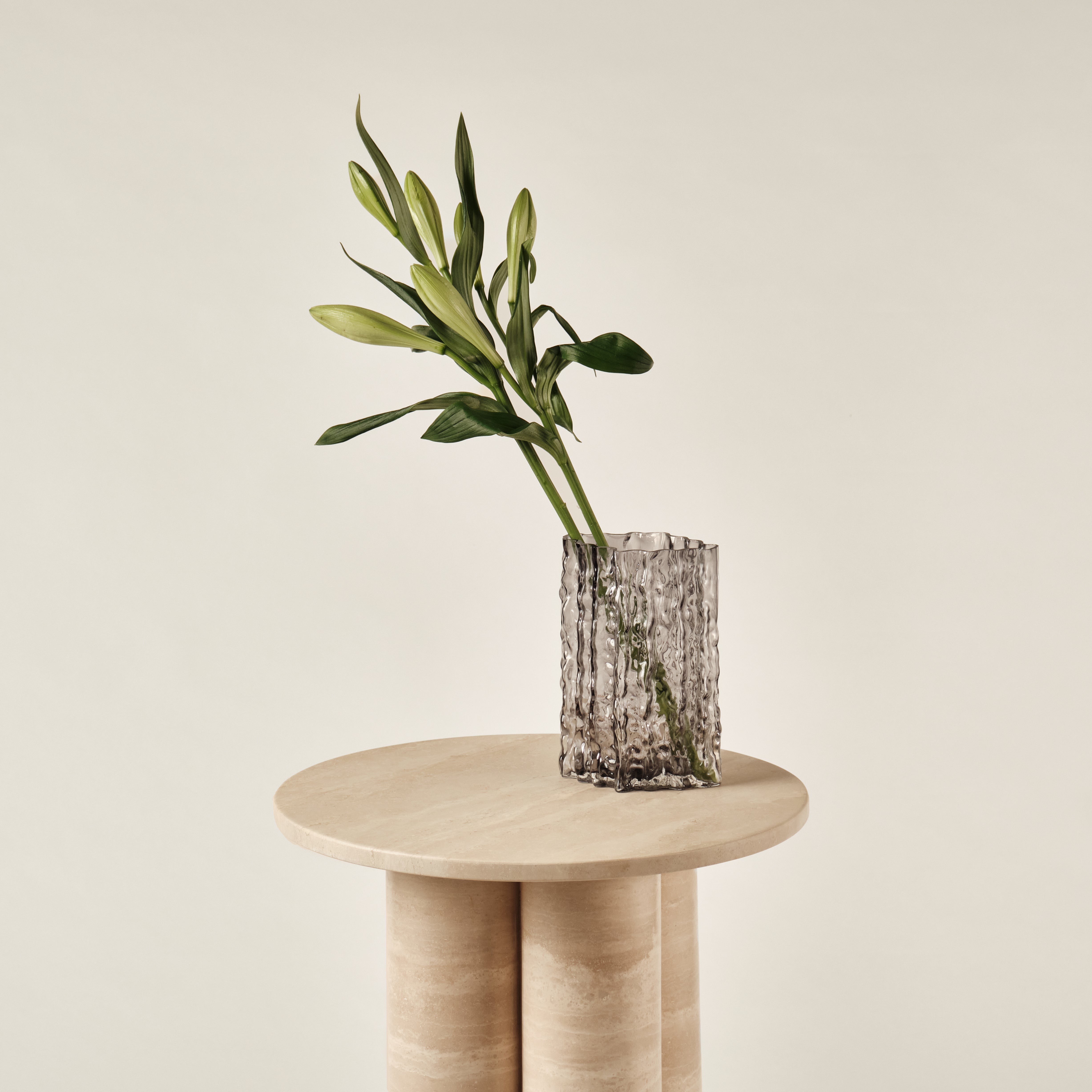 Beistelltisch aus Travertin mit runder Tischplatte, dekoriert mit einer eleganten Glasvase in Rauchoptik und frischen grünen Blumen. Perfekte Kombination aus natürlichem Stein und modernem Dekor für zeitgemäße Einrichtung.