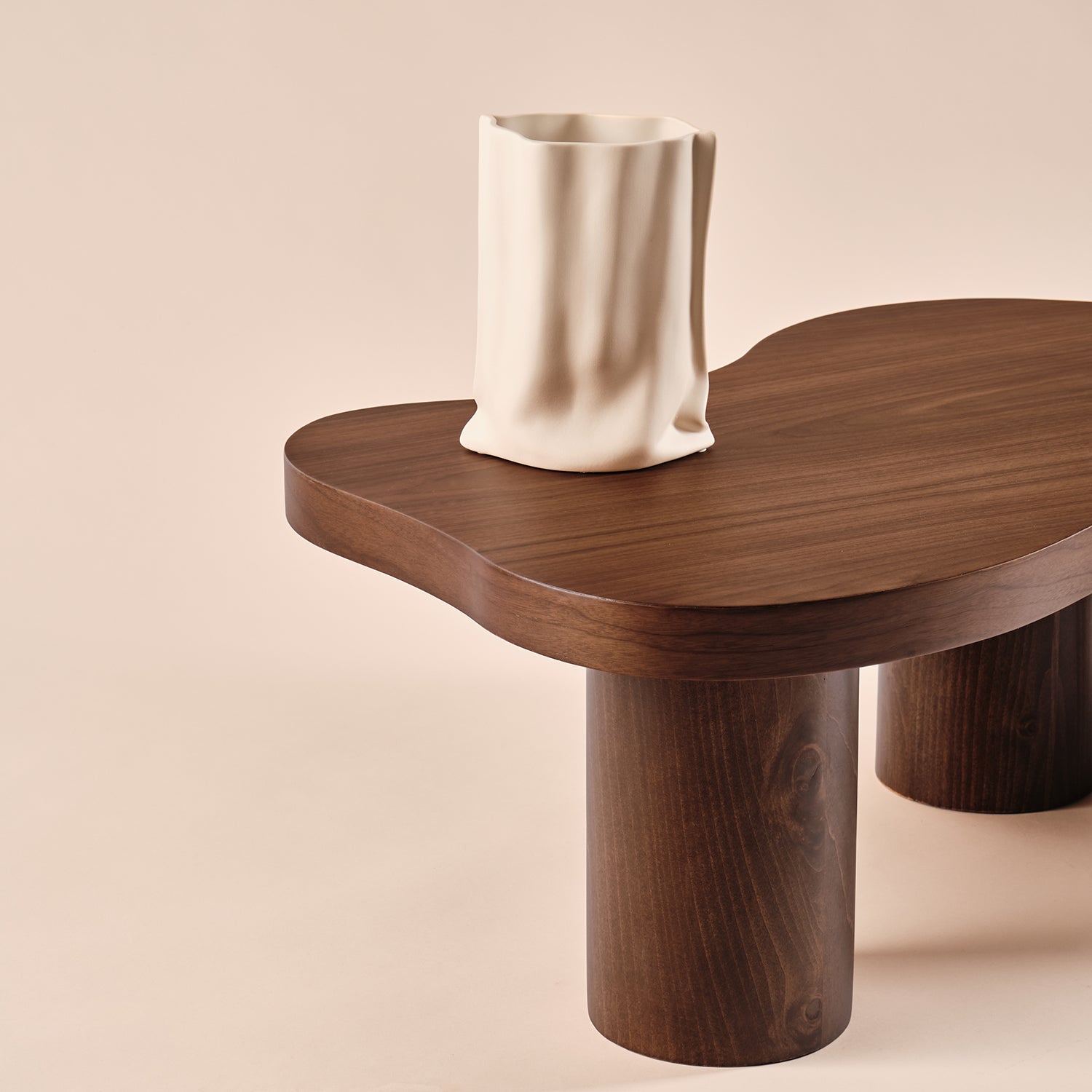 Der Couchtisch Sofie aus Holz mit einzigartiger, geschwungener Tischplatte und robusten zylindrischen Beinen. Auf der Tischplatte steht eine stilvolle Keramikvase mit skulpturaler Optik, die den minimalistischen und modernen Look des Designs unterstreicht.