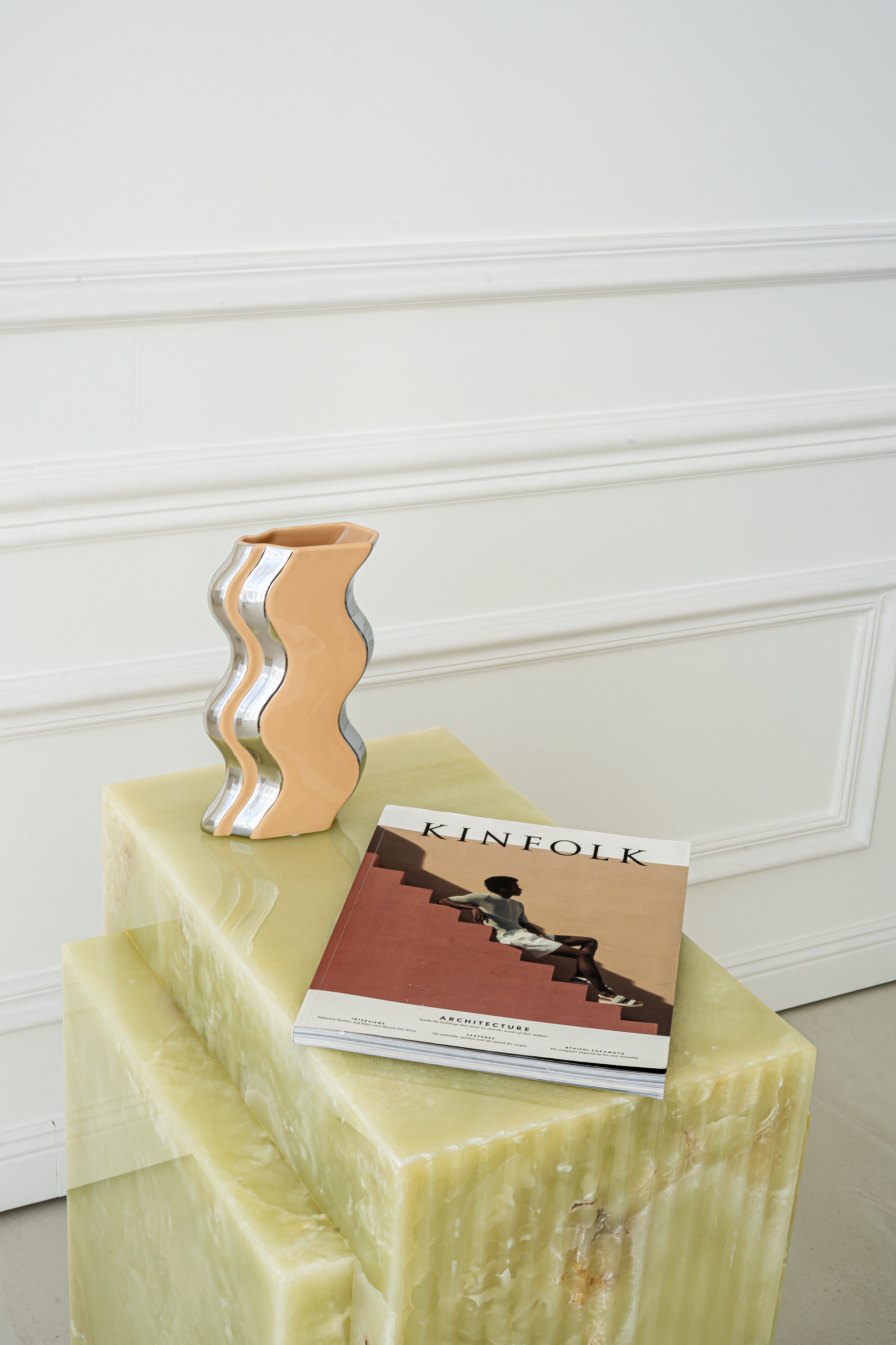 Beistelltisch Eden aus grünem Onyx, kombiniert mit einer wellenförmigen Vase in Beige und Silber sowie einer Kinfolk-Zeitschrift, perfekt inszeniert vor einer weißen Wandverkleidung.