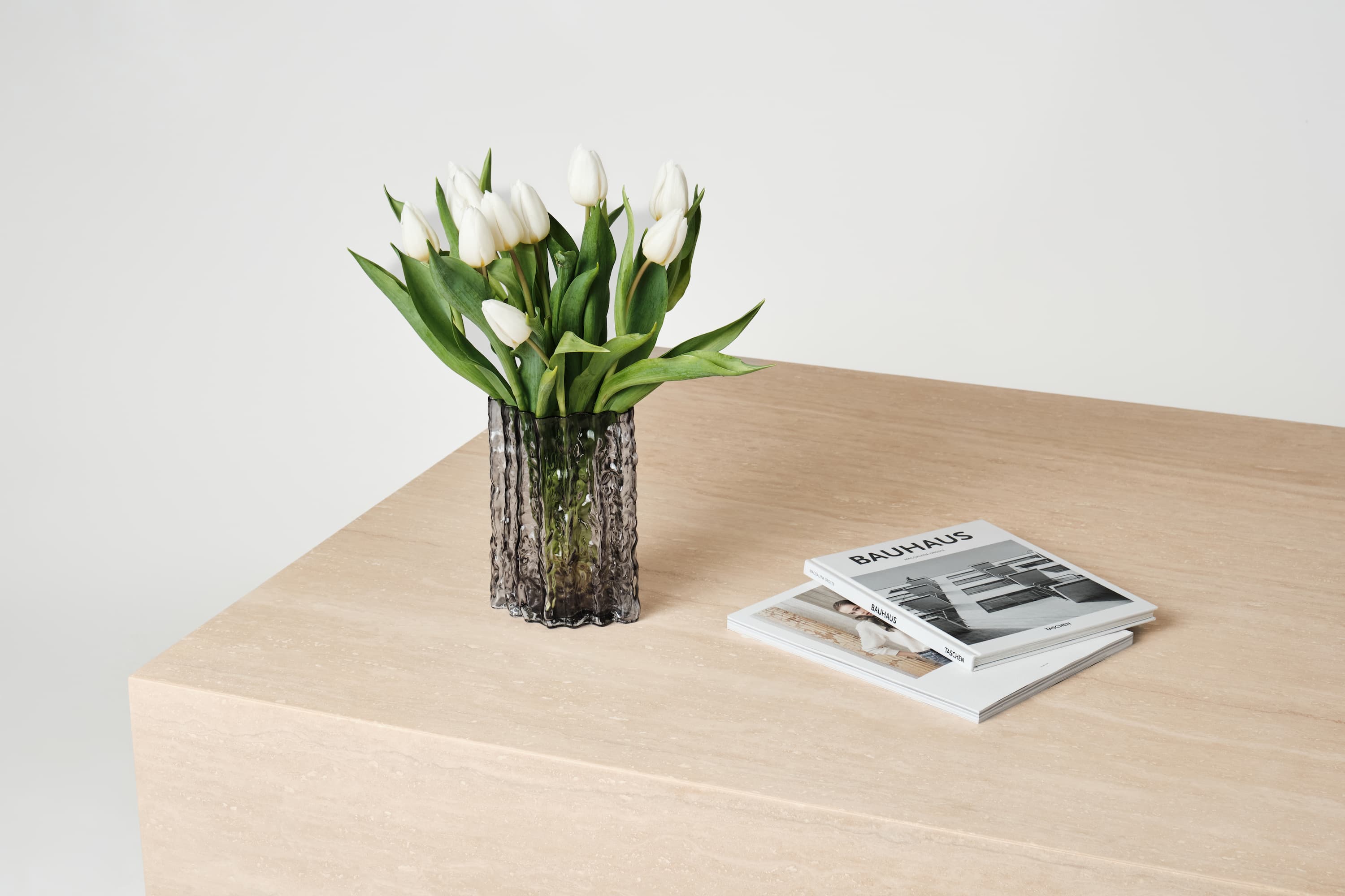 Couchtisch Gina aus edlem Travertin, dekoriert mit einem Glasgefäß mit weißen Tulpen und gestapelten Designbüchern, die eine moderne und minimalistische Ästhetik betonen.