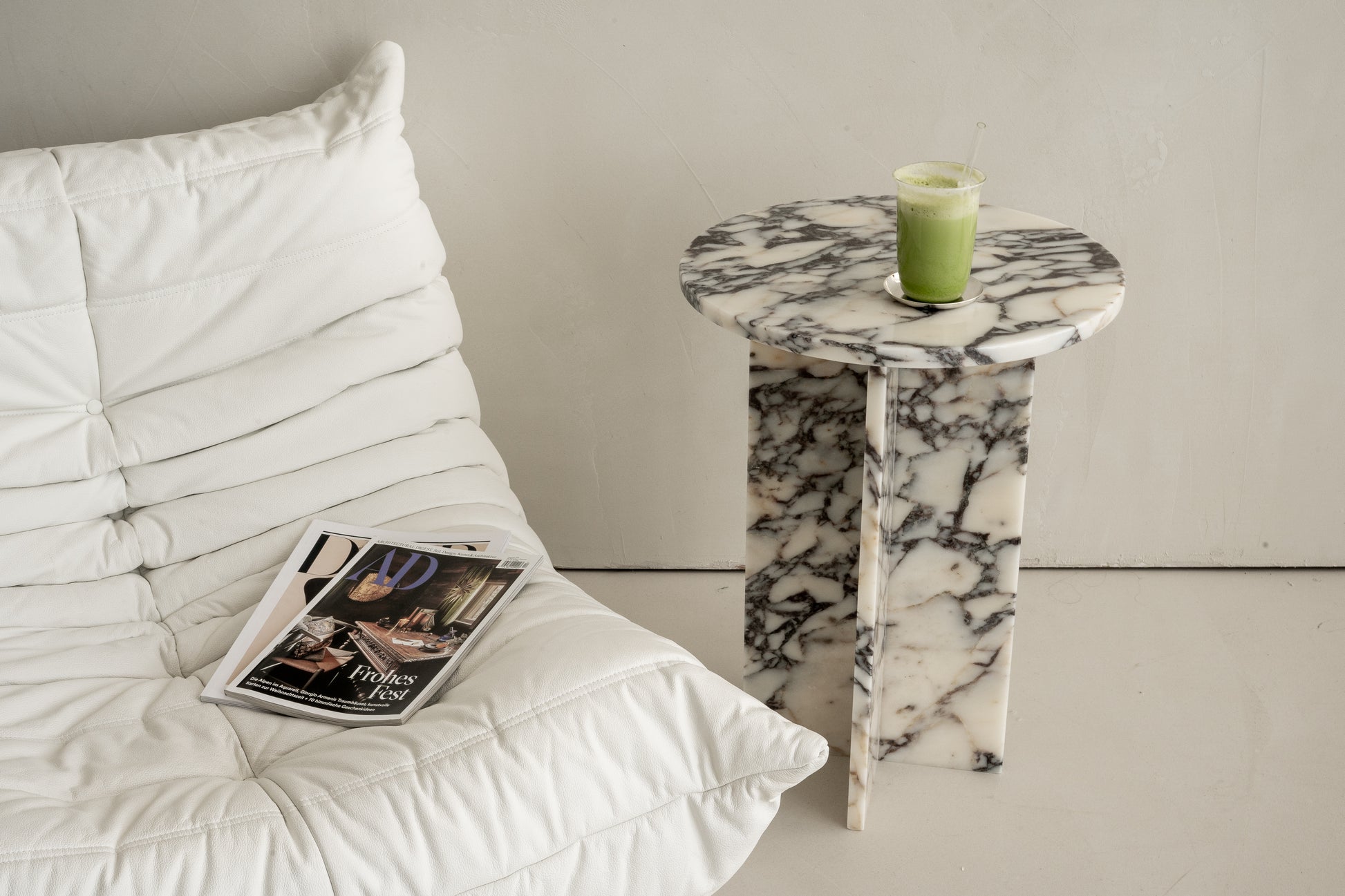 Beistelltisch aus edlem Marmor mit Schwarz-Weiß-Maserung, dekoriert mit einem Glas Matcha-Getränk, neben einem gemütlichen weißen Lounge-Sessel mit Magazin. Perfekt für moderne und elegante Wohnraumgestaltungen.