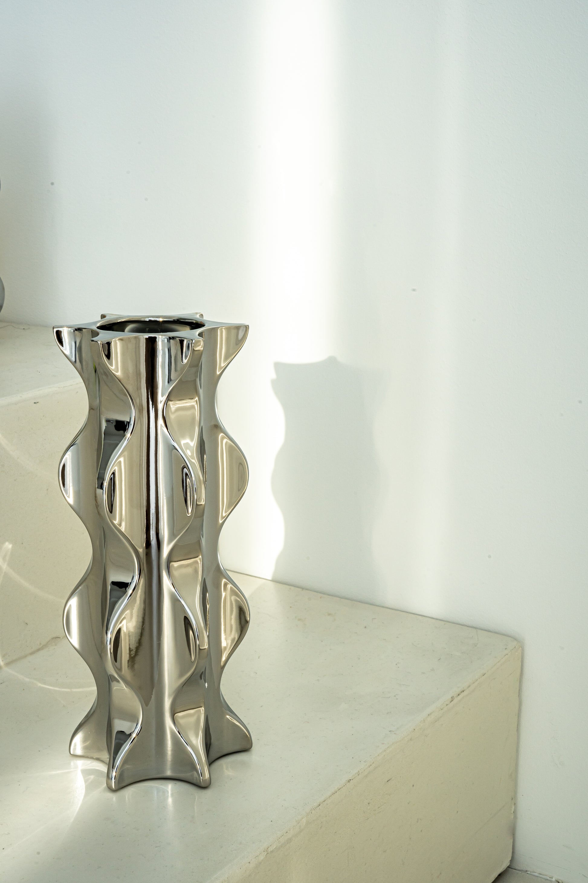 Die Porzellanvase "Cactus" aus der Kollektion in einem hochglänzenden Chrom-Finish steht dekorativ auf minimalistischen Treppenstufen. Die wellenförmige Silhouette und die reflektierende Oberfläche verleihen dem Raum eine moderne Eleganz und stilvolle Akzente.