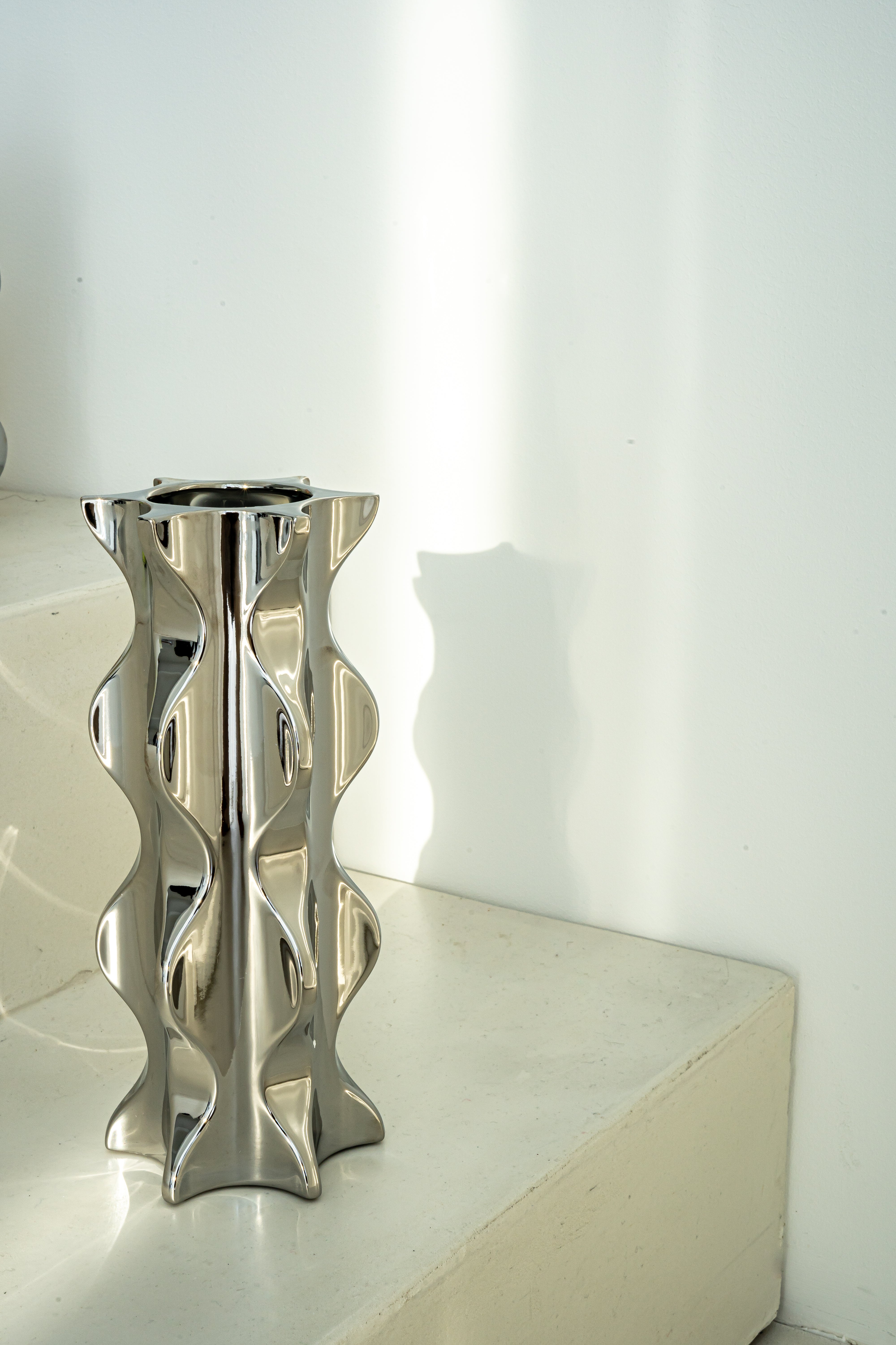 Die Porzellanvase "Cactus" aus der Kollektion in einem hochglänzenden Chrom-Finish steht dekorativ auf minimalistischen Treppenstufen. Die wellenförmige Silhouette und die reflektierende Oberfläche verleihen dem Raum eine moderne Eleganz und stilvolle Akzente.