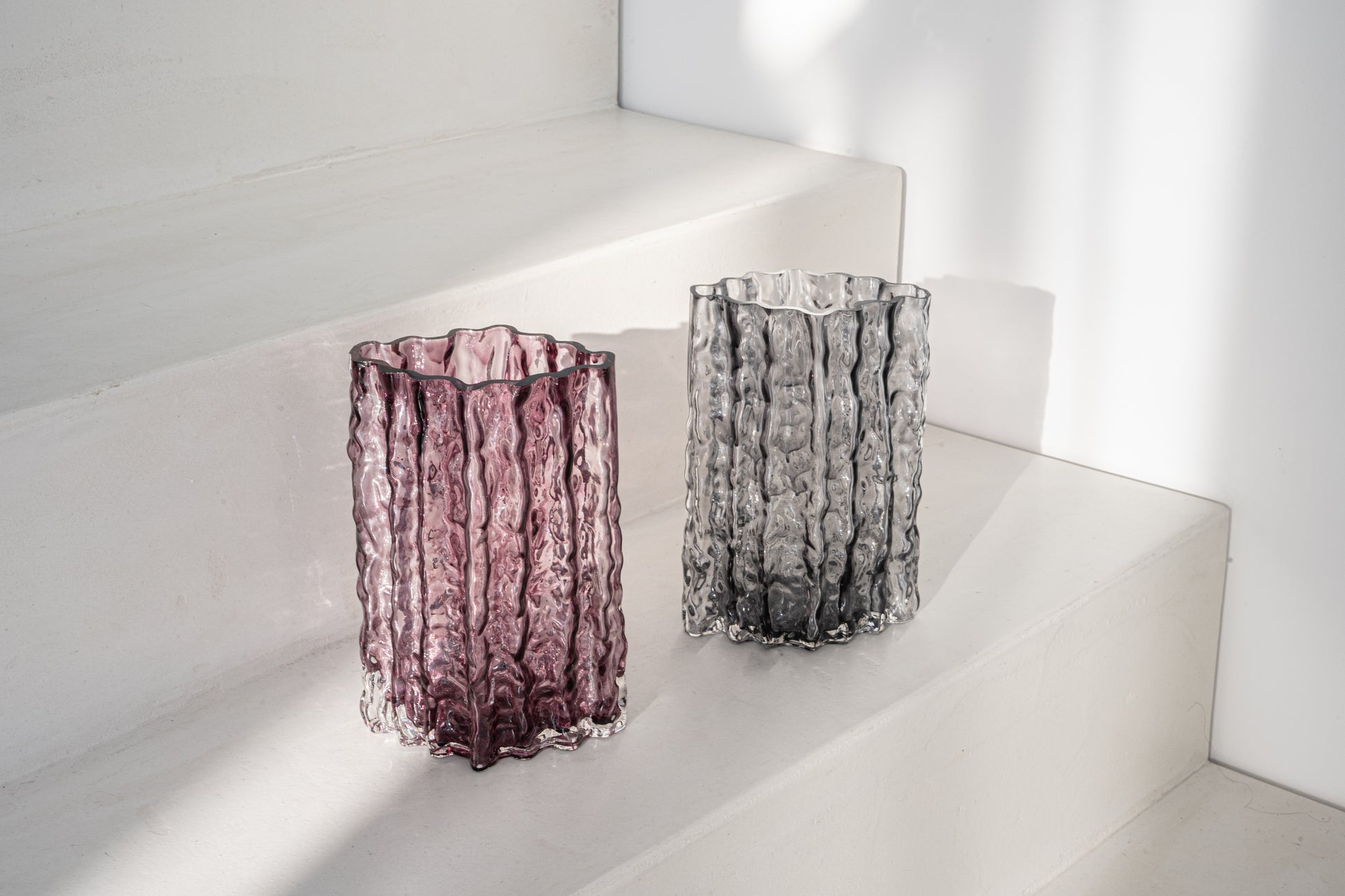 Zwei Glasvasen der Kollektion Raw, eine in kräftigem Violett und die andere in elegantem Grau, mit wellenförmiger Struktur, stilvoll arrangiert auf einer weißen Treppenstufe im Tageslicht.