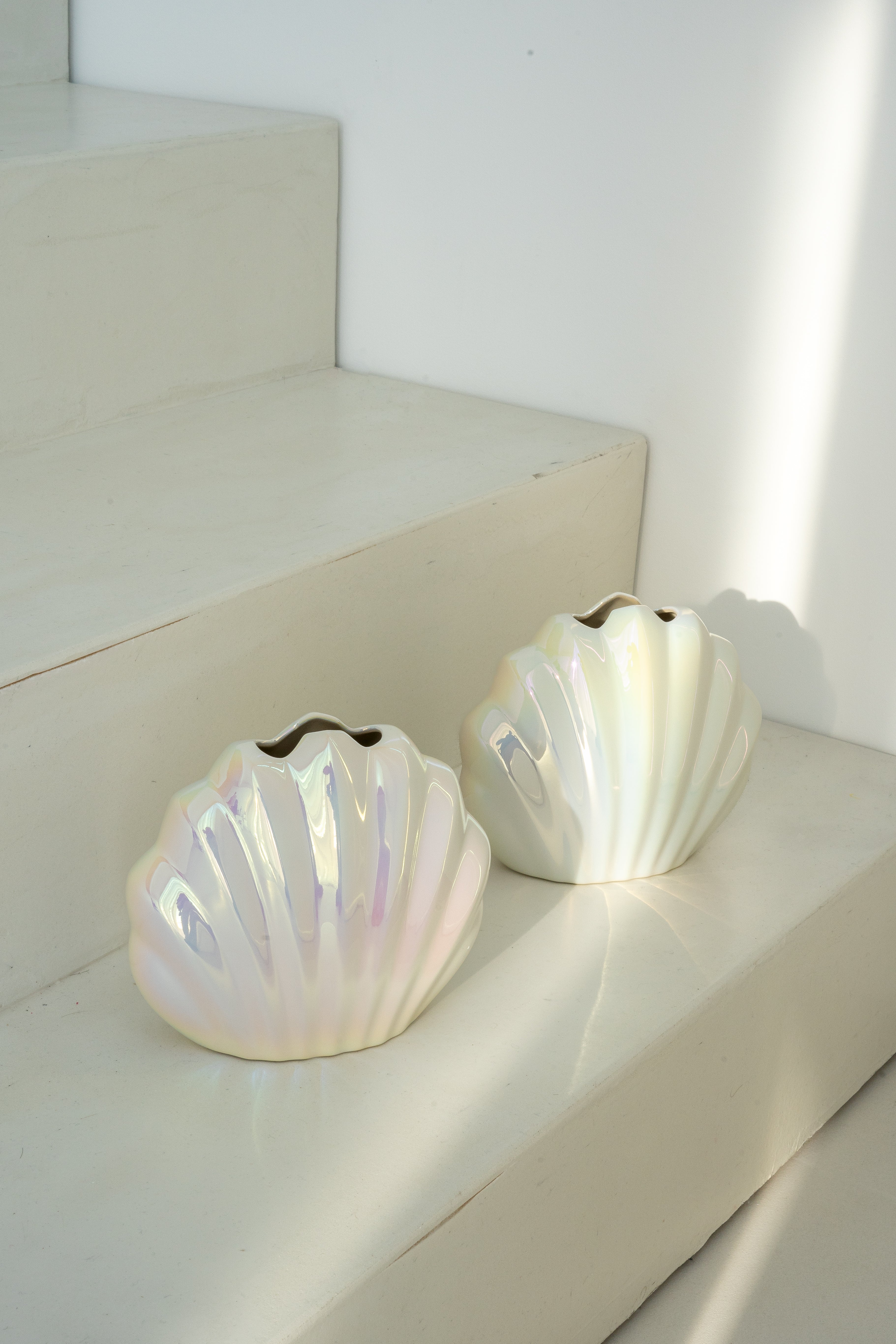 Zwei Porzellanvasen "Seashell Rainbow" in Form stilisierter Muscheln mit schimmernder Regenbogen-Glasur, elegant auf weißen Treppenstufen arrangiert. Ideal für maritime und moderne Wohnaccessoires.