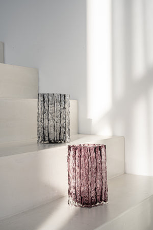 Zwei Glasvasen der Kollektion Raw, eine in Rosa und eine in Grau, aus hochwertigem Glas. Die Vasen stehen auf einer weißen Treppe und werden von natürlichem Licht akzentuiert, das das strukturierte Design hervorhebt.