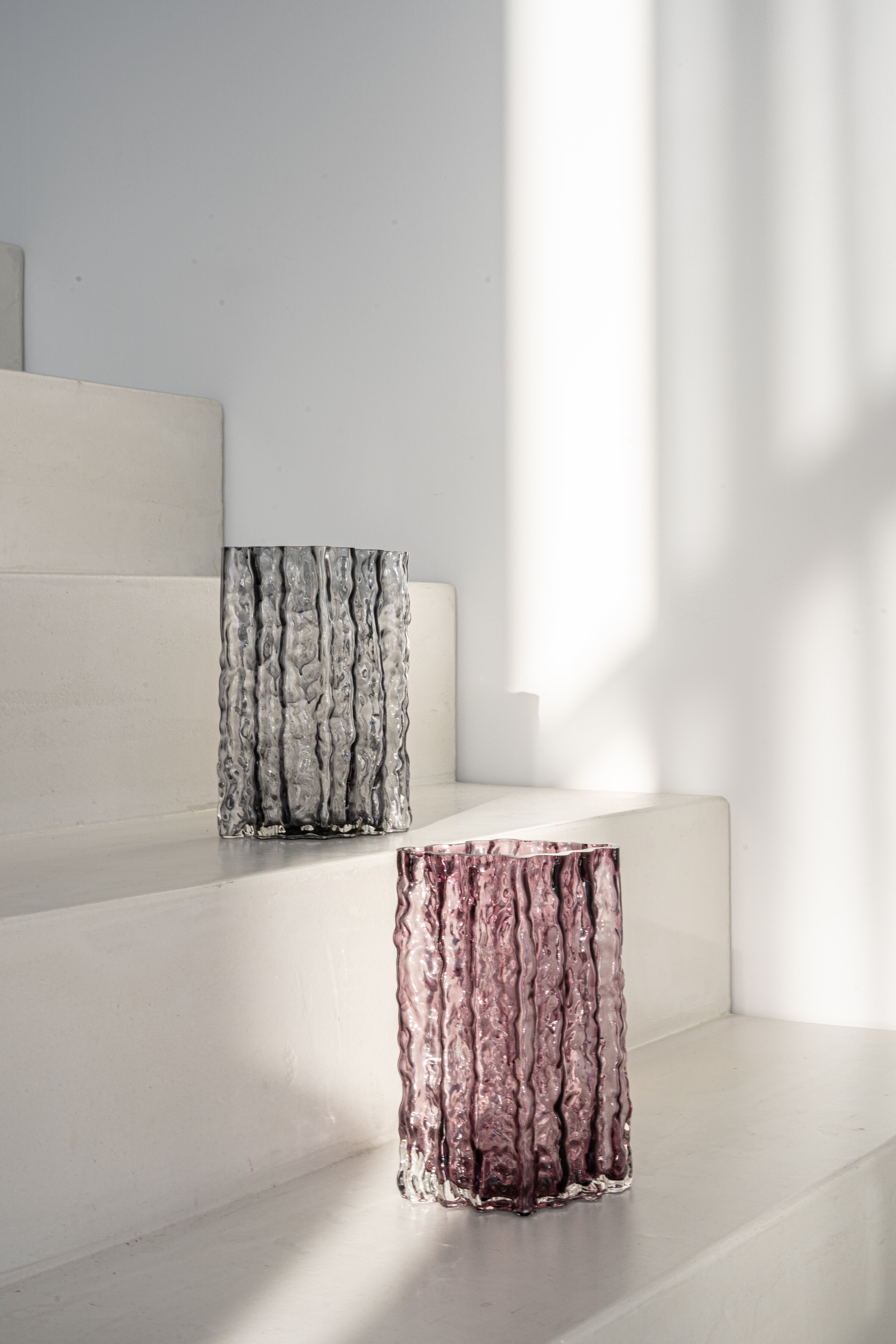 Zwei Glasvasen der Kollektion Raw, eine in Rosa und eine in Grau, aus hochwertigem Glas. Die Vasen stehen auf einer weißen Treppe und werden von natürlichem Licht akzentuiert, das das strukturierte Design hervorhebt.